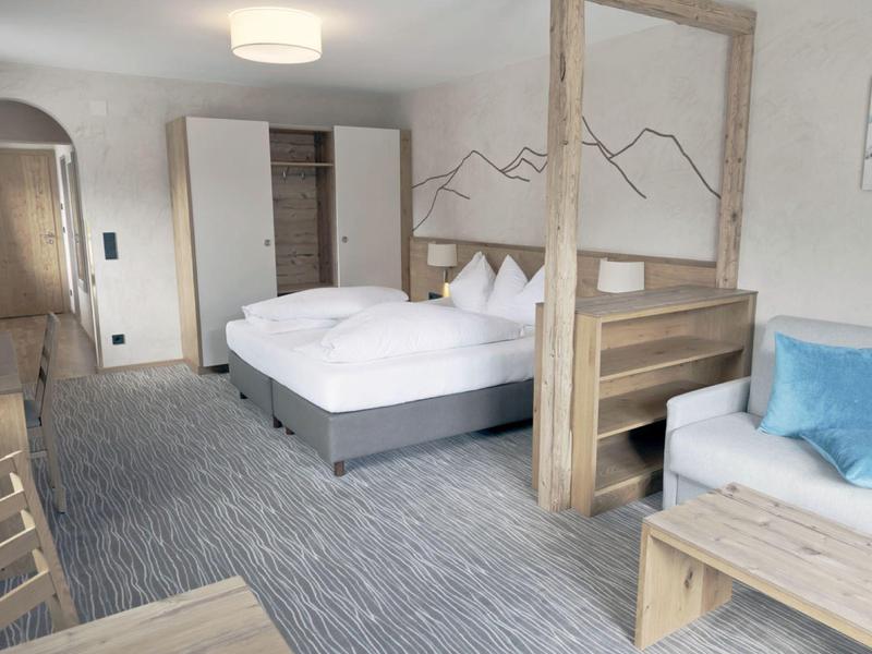 Hell eingerichtetes Schlafzimmer mit großem Bett, Couch, Holztrennwand und Teppichboden.