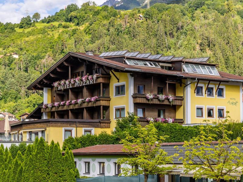 Großes gelbes Alpenhaus mit hölzernen Balkonen vor bewaldetem Hügel und blauem Himmel.