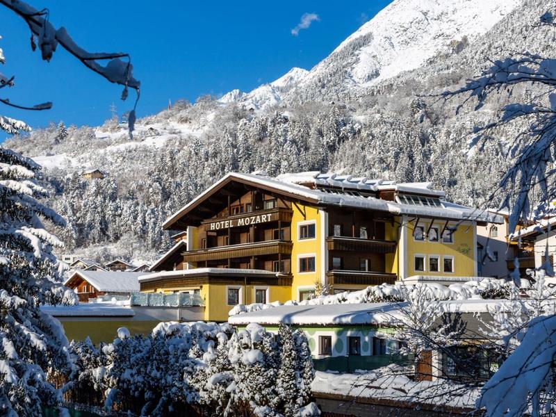 Gelbes Chalet mit Holzbalustraden, umgeben von schneebedeckten Bäumen und Bergen im Winter.