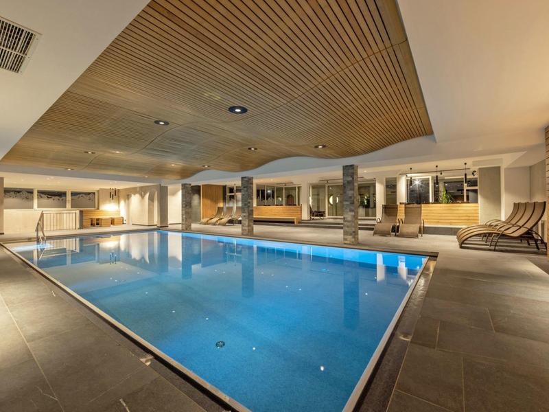 Moderner, innenliegender Swimmingpool mit Holzdecke, bequemen Liegestühlen und warmem Licht.