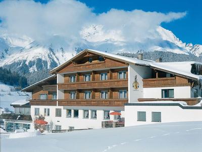 Großes Holzhaus im Schnee, umgeben von Bergen, blauer Himmel mit Wolken.