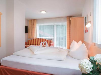 Helles Doppelzimmer mit großem Bett, Sträußen auf Nachttisch und Blick auf Balkon mit Vorhängen.