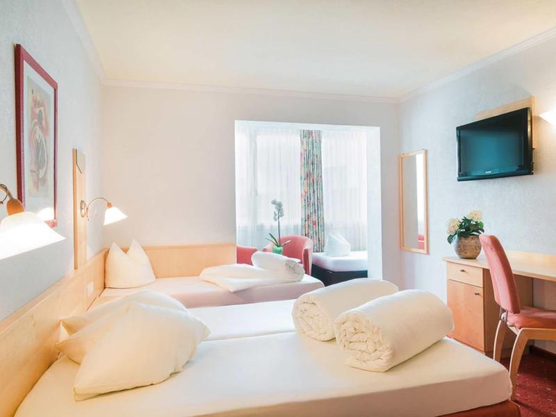 Hell beleuchtetes Hotelzimmer mit zwei Betten, Wandlampen, Schreibtisch und Fernseher an der Wand.