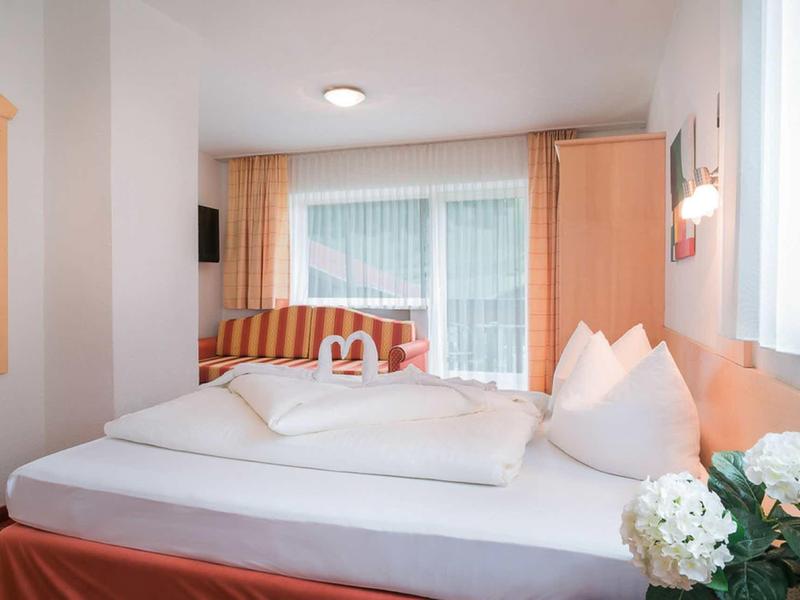 Helles Doppelzimmer mit großem Bett, Sträußen auf Nachttisch und Blick auf Balkon mit Vorhängen.