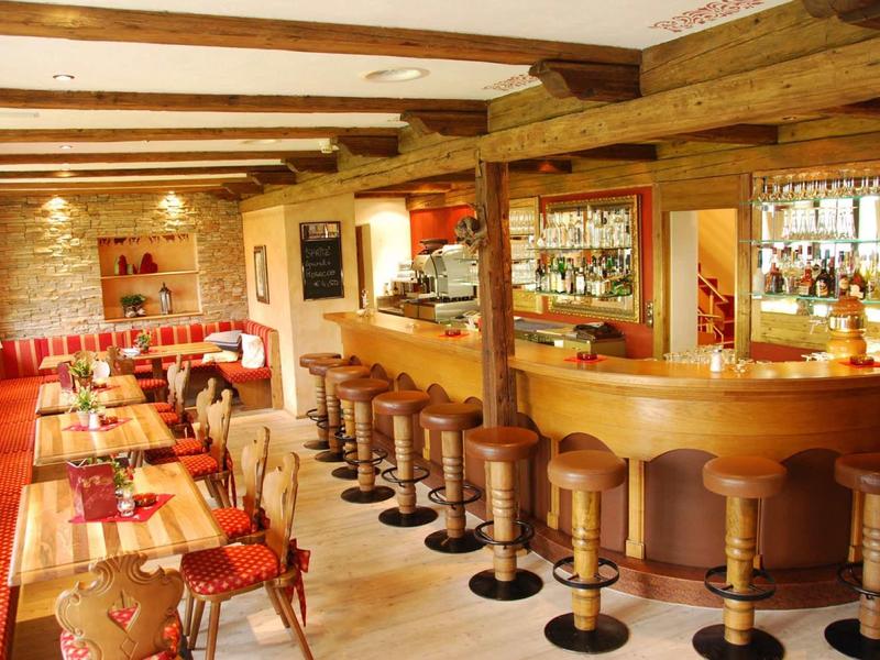 Gemütliche Bar mit Holztheke, hohen Hockern und langen Tischen mit rot-gemusterten Tischdecken.