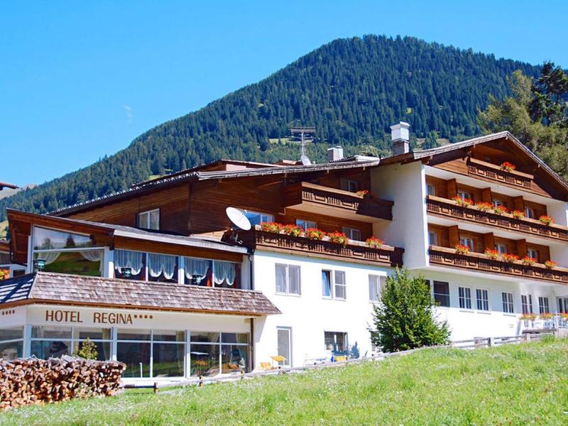 Großes Berghotel mit Balkon und Terrasse vor grünem Hügel unter blauem Himmel.