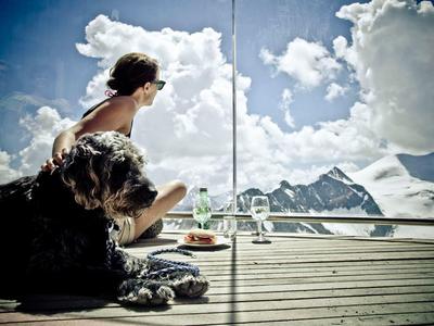 Person mit Hund sitzen auf Holzdeck, blicken auf verschneite Berge und bewölkten Himmel.