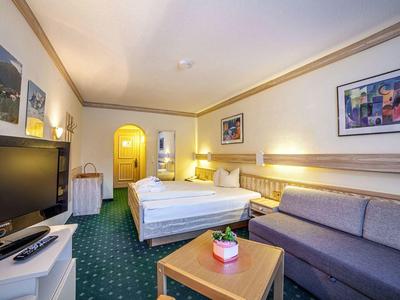 Helles Hotelzimmer mit Doppelbett, Sofa, Fernseher und grünem Teppichboden.