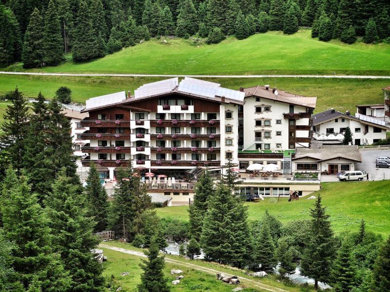 Mehrstöckiges Hotel umgeben von grünen Wiesen und dichten Tannen in einer Berglandschaft.