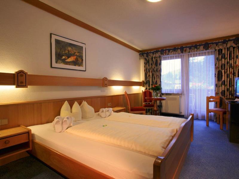 Hell eingerichtetes Hotelzimmer mit großem Doppelbett, Balkon und Fernseher.