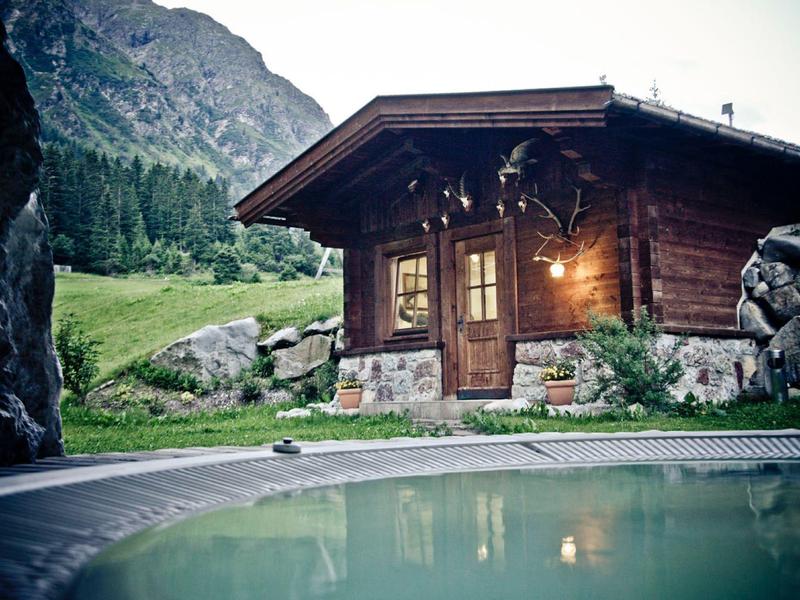Holzhütte mit Licht an, Bergwiese und Pool im Vordergrund, umgeben von Felsen und Wald.