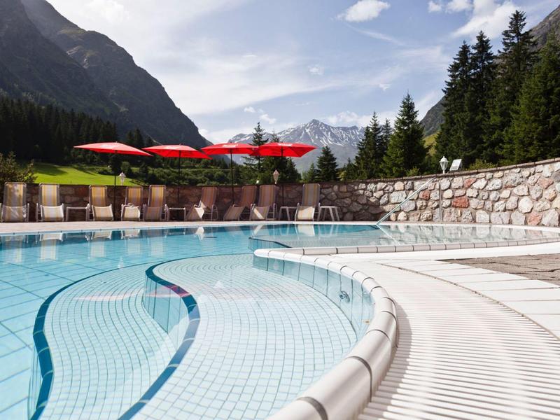 Außenpool mit klar blauem Wasser, rotem Sonnenschirm, umgeben von Bergen und grünen Bäumen.