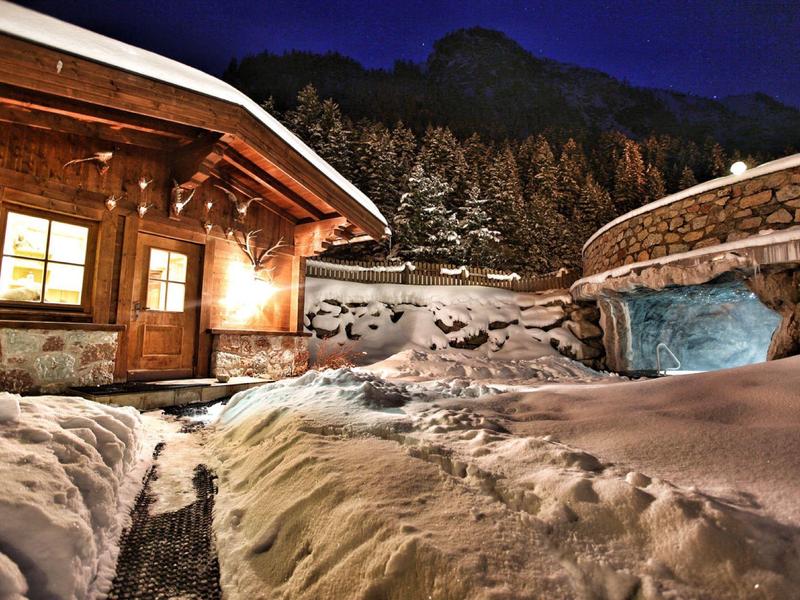 Winterlicher verschneiter Hof mit beleuchtetem Holzhaus und rundem Steingebäude bei Nacht.