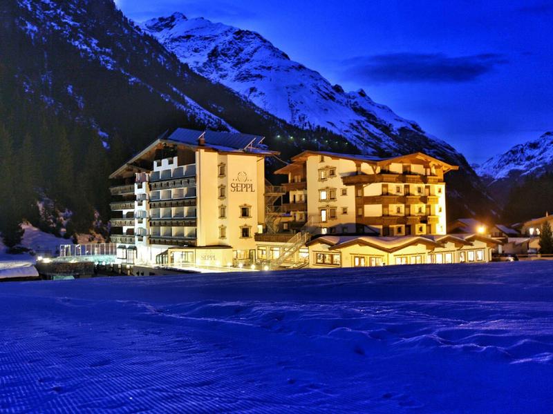 Beleuchtetes Hotel am verschneiten See mit schneebedeckten Bergen im Hintergrund bei Nacht.