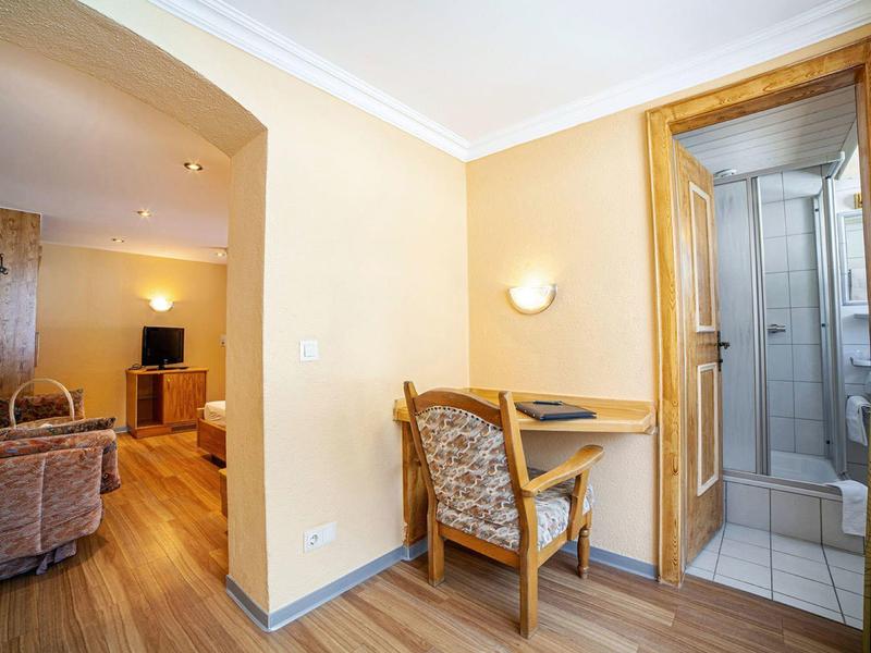 Heller Raum mit Holzboden, Sessel, Schreibtisch und angrenzendem Badezimmer mit Glastür.