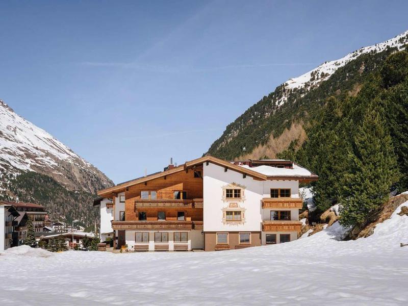 Hotel in snowy mountain landscape with green fir trees under a blue sky.