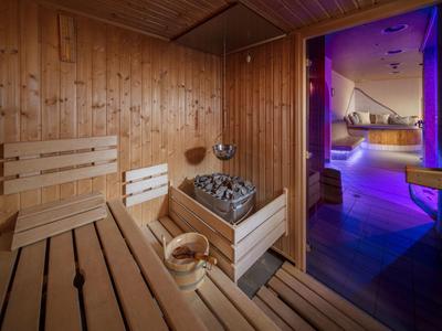 Holzsauna mit Holzbänken, Saunaofen und Eimer, daneben moderner Raum mit violettem Licht.