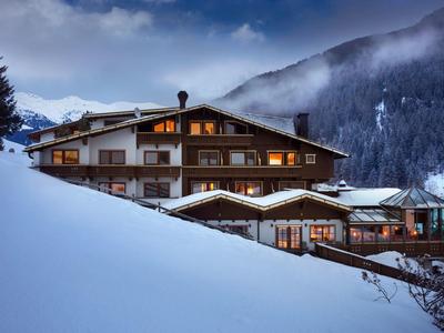 Alpenhaus mit hell erleuchteten Fenstern, umgeben von verschneiten Bergen und dunklem Abendhimmel.