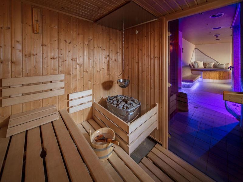 Holzsauna mit Holzbänken, Saunaofen und Eimer, daneben moderner Raum mit violettem Licht.