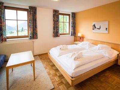 Zimmer mit Doppelbett, Holzboden, zwei Fenstern, Tisch und bunten Vorhängen, Wänden in Gelb.