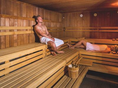 Zwei Männer entspannen in einer holzvertäfelten Sauna mit mehrstufigen Bänken und Sauna-Eimer.