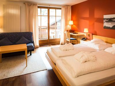 Helles Schlafzimmer mit Doppelbett, Sofa, Holzmöbeln, roter Wand und großem Fenster mit Balkonblick.