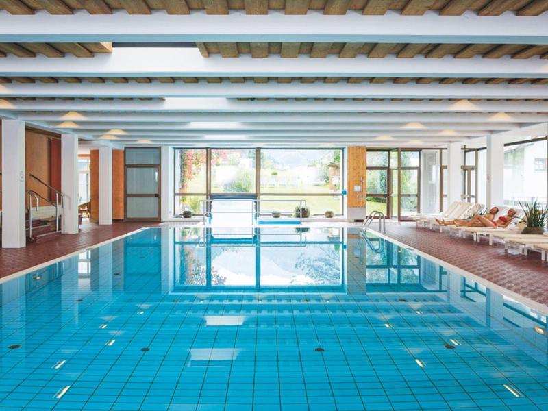 Hallenbad mit blauem Wasser, weißem Deckenraster und Liegestühlen entlang der Seitenwand.