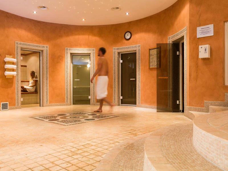 Ein runder Wellnessraum mit beige Fliesen, drei Kabinen und einer Person in einem weißen Handtuch.