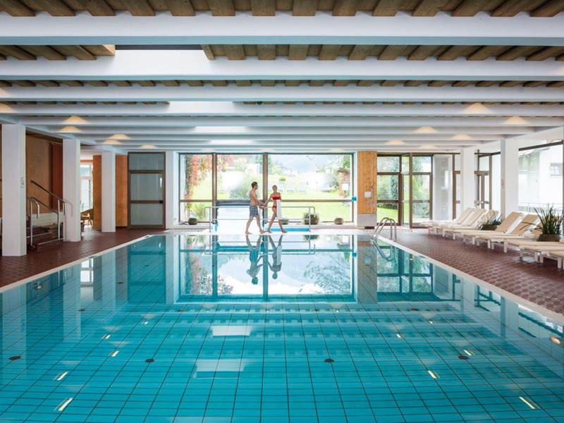 Innen-Schwimmbad mit klarem Wasser, Holzfußboden und weißen Liegestühlen am Beckenrand.