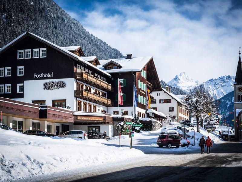 Schneebedecktes Dorf mit Hotel, Autos und Kirche vor bewölkten Bergen in klarer Winterlandschaft.