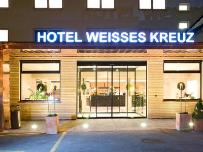 Fassade eines Hotels mit beleuchtetem blauen Schild "HOTEL WEISSES KREUZ" und Glas-Eingangstür.