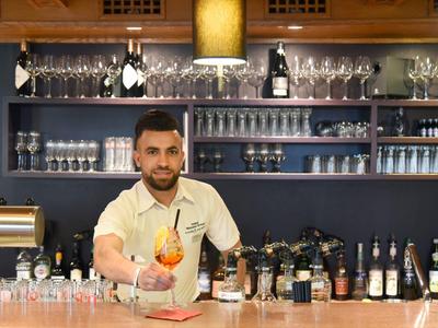 Barkeeper reicht ein buntes Cocktailglas über eine Bar mit Flaschen und Gläsern im Hintergrund.