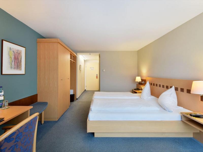 Helles Hotelzimmer mit Doppelbett, blauer Wand, Holzmöbeln, Schreibtisch und gemütlicher Beleuchtung.