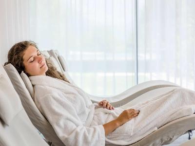 Eine junge Frau entspannt in einem weißen Bademantel auf einem bequemen Liegestuhl vor hellen Gardinen.