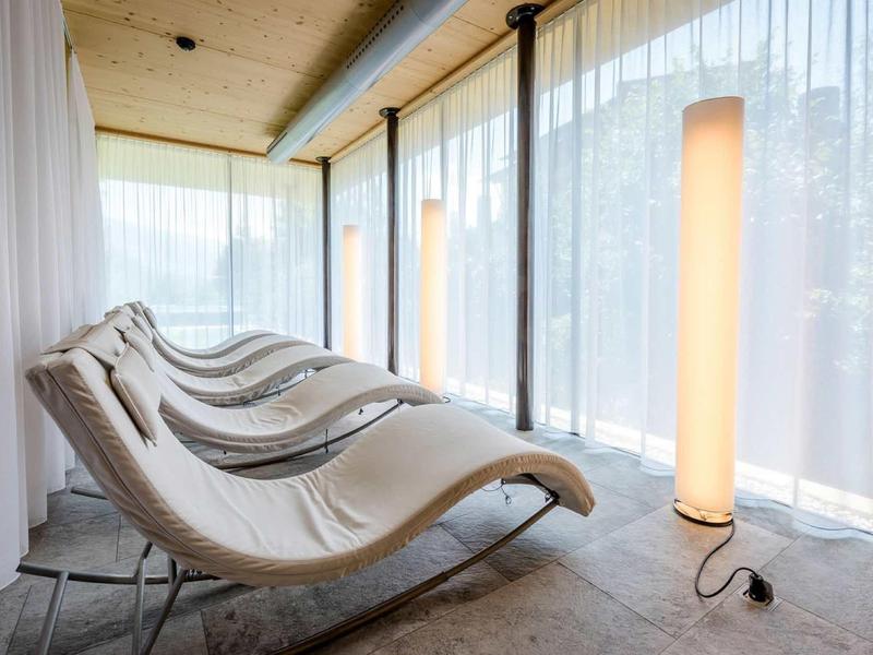 Ruheliege mit weißem Stoffbezug und moderner Stehlampe in hellem Raum mit großen Fenstern.