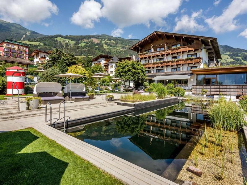Luxuriöses Hotel mit Holzdetails, grüner Gartenanlage und klarem Teich vor Bergkulisse bei Sonne.