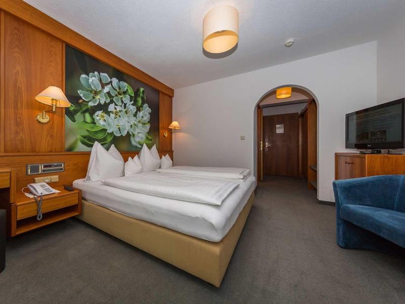 Gemütliches Hotelzimmer mit Doppelbett, Holzdekor, blauem Sessel und Fernseher an der Wand.