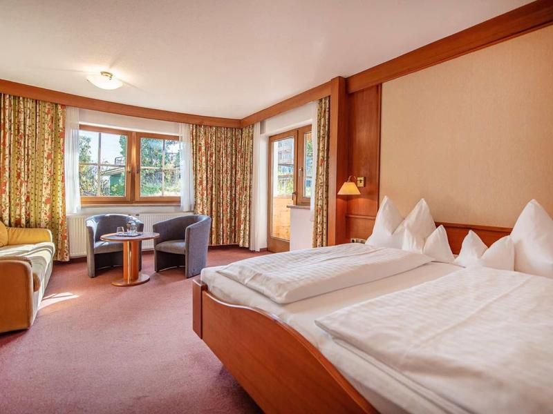 Gemütliches Schlafzimmer mit großem Bett, Sessel, Schreibtisch und Fenstern mit blickdichten Vorhängen.