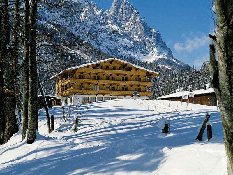 Holzhaus in verschneitem Tal vor schneebedeckten Bergen unter klarem blauem Himmel.