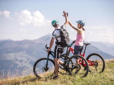 Two cyclists wearing helmets and backpacks high-five on a hill with mountains in the background.
