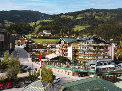 Hotel mit Holzfassade und Balkon in einem grünen Bergtal unter blauem Himmel.