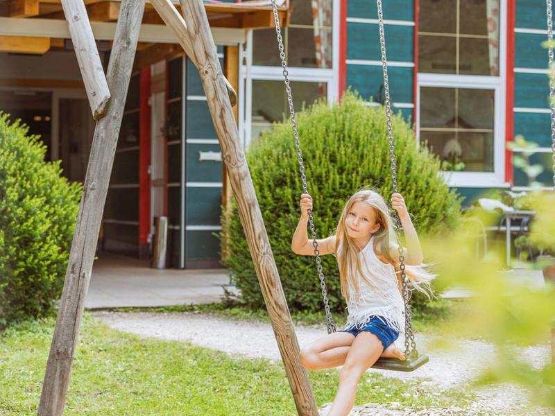Mädchen schaukelt auf einem Holzschaukelgestell vor einem Hotel mit grüner Fassade.