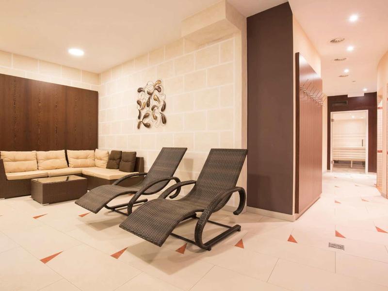 Moderner Wartebereich mit beiger Eckcouch, zwei schwarzen Liegestühlen und heller, gefliester Wand.