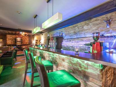 Nowoczesny hotelowy bar z wysokimi stołami, zielonymi stołkami barowymi i stylowym oświetleniem na kamiennej ścianie.