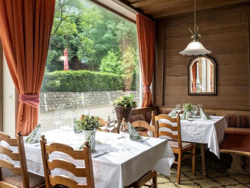 Gemütliches Esszimmer mit Holzmöbeln, weißen Tischdecken, Pflanzen und großen Fenstern ins Grüne.