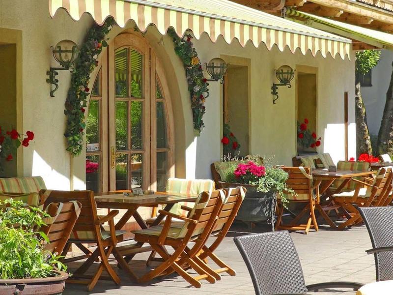 Gemütliche Außenterrasse mit Holztischen, Stühlen und bunten Blumen an einer gelben Hauswand.