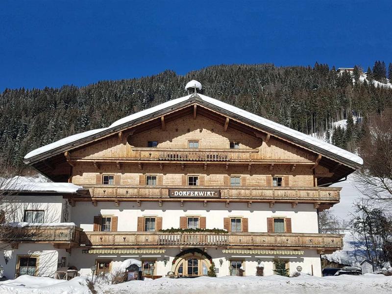 Großes Holzhaus mit Balkon im Schnee, hinter dem Haus bewaldeter Hügel unter blauem Himmel.