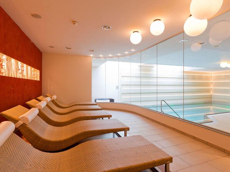 Ruhebereich mit Liegestühlen neben verglaster Wand mit Blick auf Schwimmbadbereich.