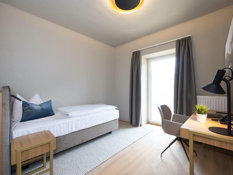 Helles, modernes Hotelzimmer mit Einzelbett, Schreibtisch und Fenster mit Vorhängen.