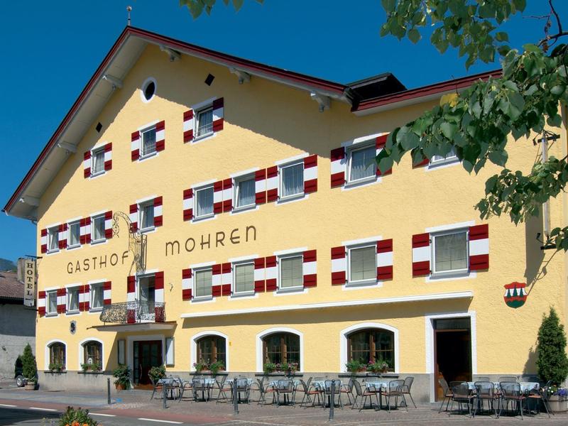 Gelbes Gasthaus mit roten und weißen Fensterläden und Außensitzbereich unter blauem Himmel.